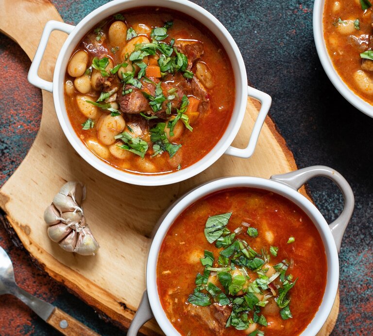 Serbische Bohnensuppe in drei runden Schalen aus Porzellan / Holzbrett / frischer Knoblauch / Suppenlöffel / Draufsicht | © Shutterstock / Daria Mladenovic
