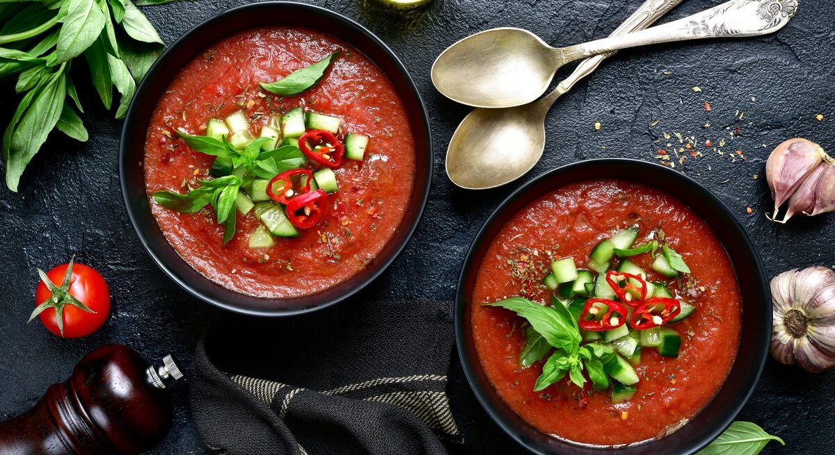 Zwei runde Schalen mit der spanischen Gemüsesuppe Gazpacho Andaluz / zwei Löffel / frischer Knoblauch / Kräuter / Pfeffermühle /Tuch / eine Tomate / Draufsicht