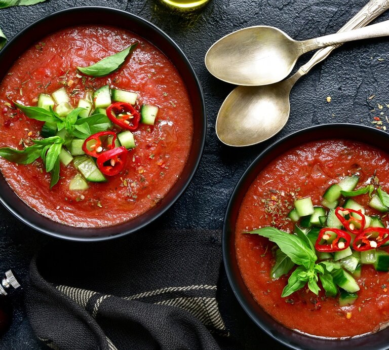 Zwei runde Schalen mit der spanischen Gemüsesuppe Gazpacho Andaluz / zwei Löffel / frischer Knoblauch / Kräuter / Pfeffermühle /Tuch / eine Tomate / Draufsicht | © Shutterstock / Liliya Kandrashevich