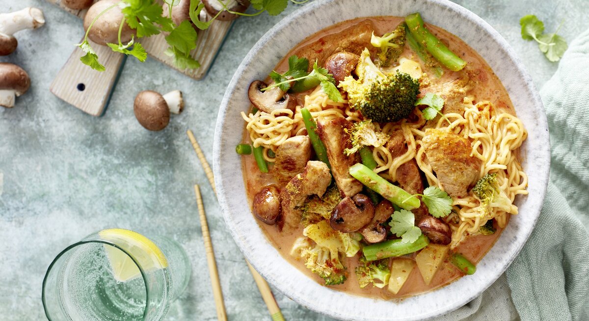 Ein helle Teller mit dem Thai-Curry auf einem hellblauem Untergrund mit grünem Gemüse und Pilzen daneben. Alles direkt von oben fotografiert.