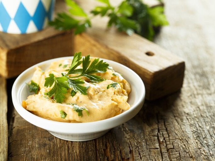 Weißer Teller mit Obazda, garniert mit Petersilie, auf einem rustikalen Holztisch, im Hintergrund ein bayerisches Muster. | © iStock/ Mariha-kitchen