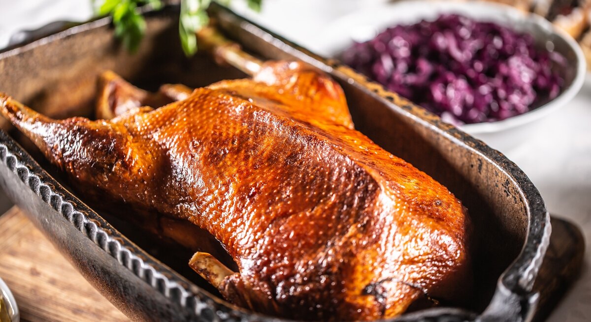Eine goldbraune gebackene Gans in einer tiefen Backform auf einem Holzschneidebrett und hellem Küchentisch. Im Hintergrund sind unscharf Rotkohl und die Brezeln zu erkennen.