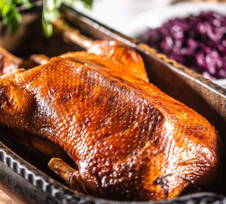 Eine goldbraune gebackene Gans in einer tiefen Backform auf einem Holzschneidebrett und hellem Küchentisch. Im Hintergrund sind unscharf Rotkohl und die Brezeln zu erkennen. | © Shutterstock/ Marian Weyo