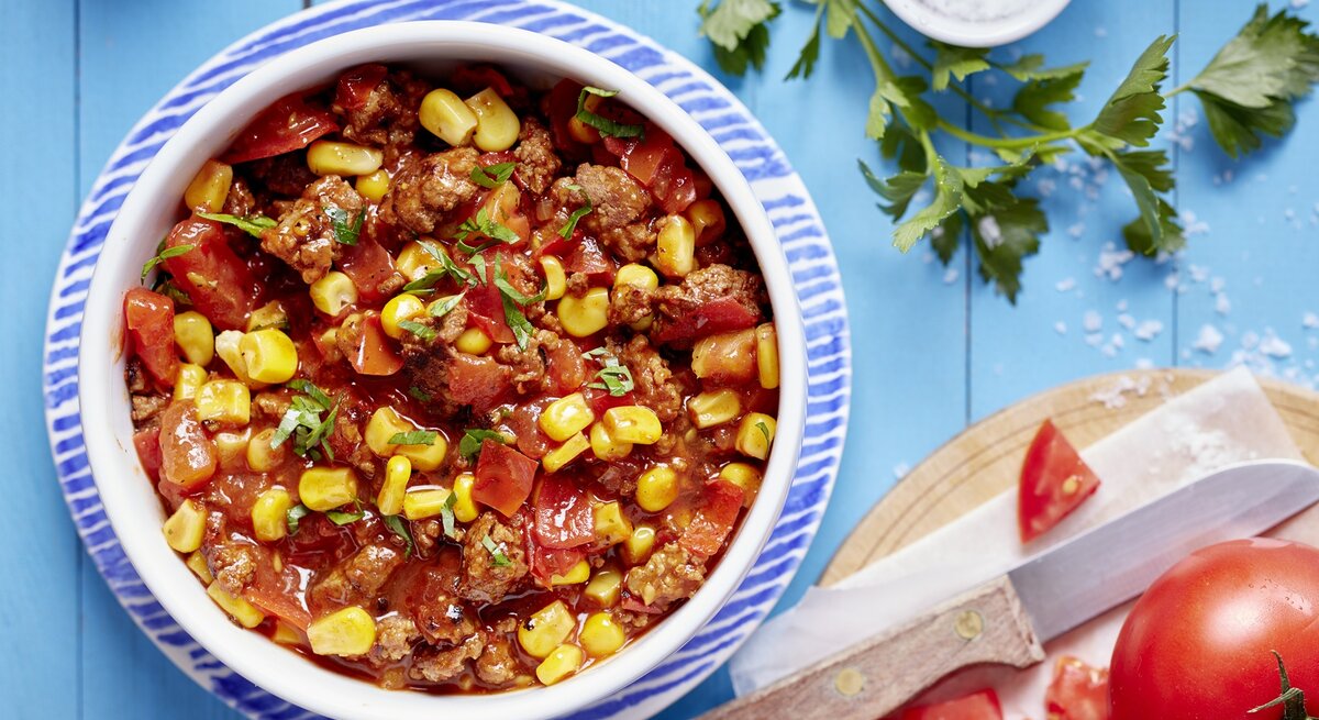 Eine Schale Chili con Carne in einem weiß-blauen Teller auf dem blauen Tisch. Alles direkt von oben fotografiert.