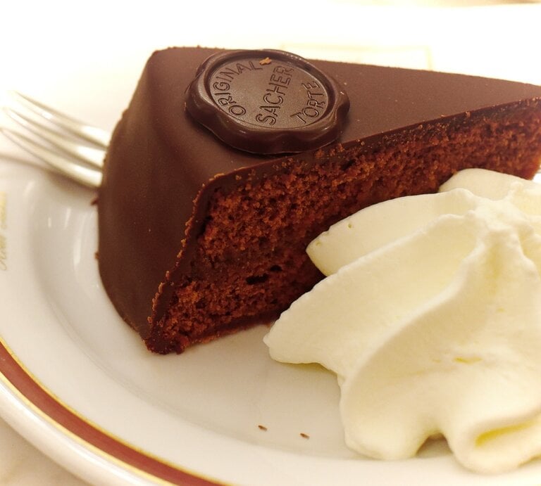 Ein Stück Sachertorte / aufgeschlagene Sahne / Kuchengabel / runder Kuchenteller / Draufsicht schräg | © Shutterstock / Margy Crane