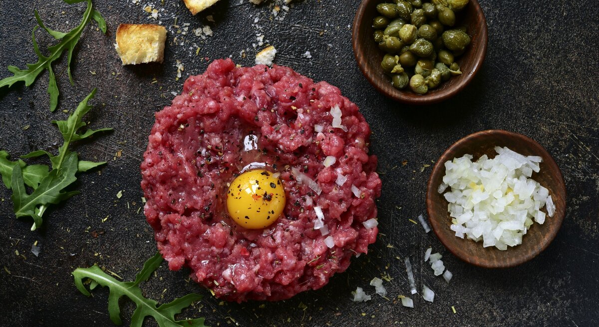 Klassisches Rintertatar / rohes, aufgeschlagenes Ei / Gewürze / schälchen mit fein geschnittenen rohen Zwiebel und kleinen Kapern / Brotwürfel und Ruccolablättern