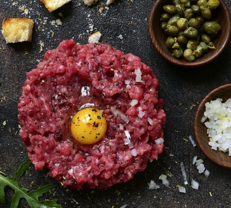 Klassisches Rintertatar / rohes, aufgeschlagenes Ei / Gewürze / schälchen mit fein geschnittenen rohen Zwiebel und kleinen Kapern / Brotwürfel und Ruccolablättern | © Getty Images/Lilechka75
