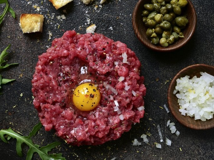 Klassisches Rintertatar / rohes, aufgeschlagenes Ei / Gewürze / schälchen mit fein geschnittenen rohen Zwiebel und kleinen Kapern / Brotwürfel und Ruccolablättern | © Getty Images/Lilechka75