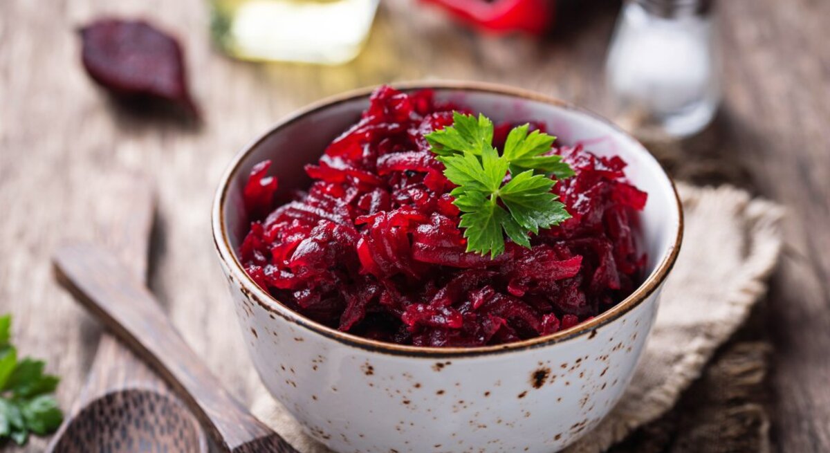 Das Bild zeigt eine Schüssel mit Rote-Bete-Salat, garniert mit einem Blatt Petersilie. Die Schüssel steht auf einem rustikalen Holztisch und im Hintergrund sieht man eine Ölflasche, eine Salzstreuer und vermutlich eine Pfeffermühle.
