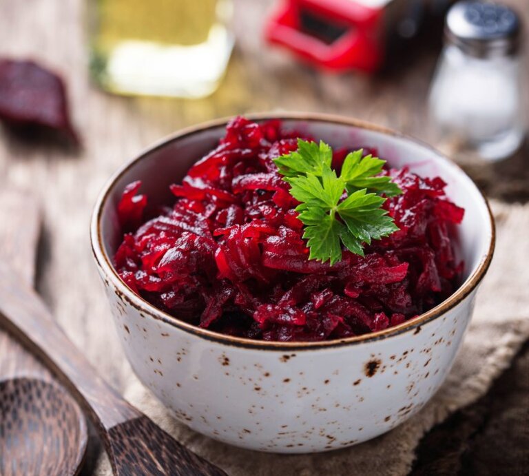 Das Bild zeigt eine Schüssel mit Rote-Bete-Salat, garniert mit einem Blatt Petersilie. Die Schüssel steht auf einem rustikalen Holztisch und im Hintergrund sieht man eine Ölflasche, eine Salzstreuer und vermutlich eine Pfeffermühle. | © AdobeStock/Yulia Furman