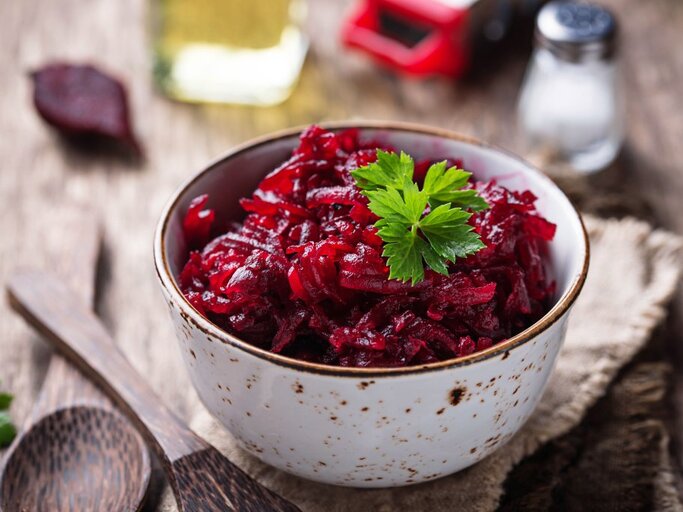 Das Bild zeigt eine Schüssel mit Rote-Bete-Salat, garniert mit einem Blatt Petersilie. Die Schüssel steht auf einem rustikalen Holztisch und im Hintergrund sieht man eine Ölflasche, eine Salzstreuer und vermutlich eine Pfeffermühle. | © AdobeStock/Yulia Furman