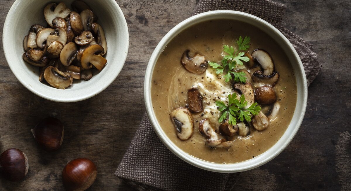 Dunkler Untergrund mit einer Schüssel der Suppe sowie frischen Champignons und Esskastanien drum herum. Alles direkt von oben fotografiert.