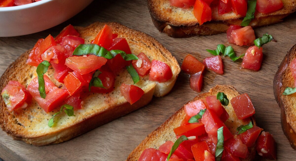 Knusprige Bruschetta mit frischen Tomatenwürfeln und Basilikumblättern auf Holzbrett neben einer weißen Schüssel.