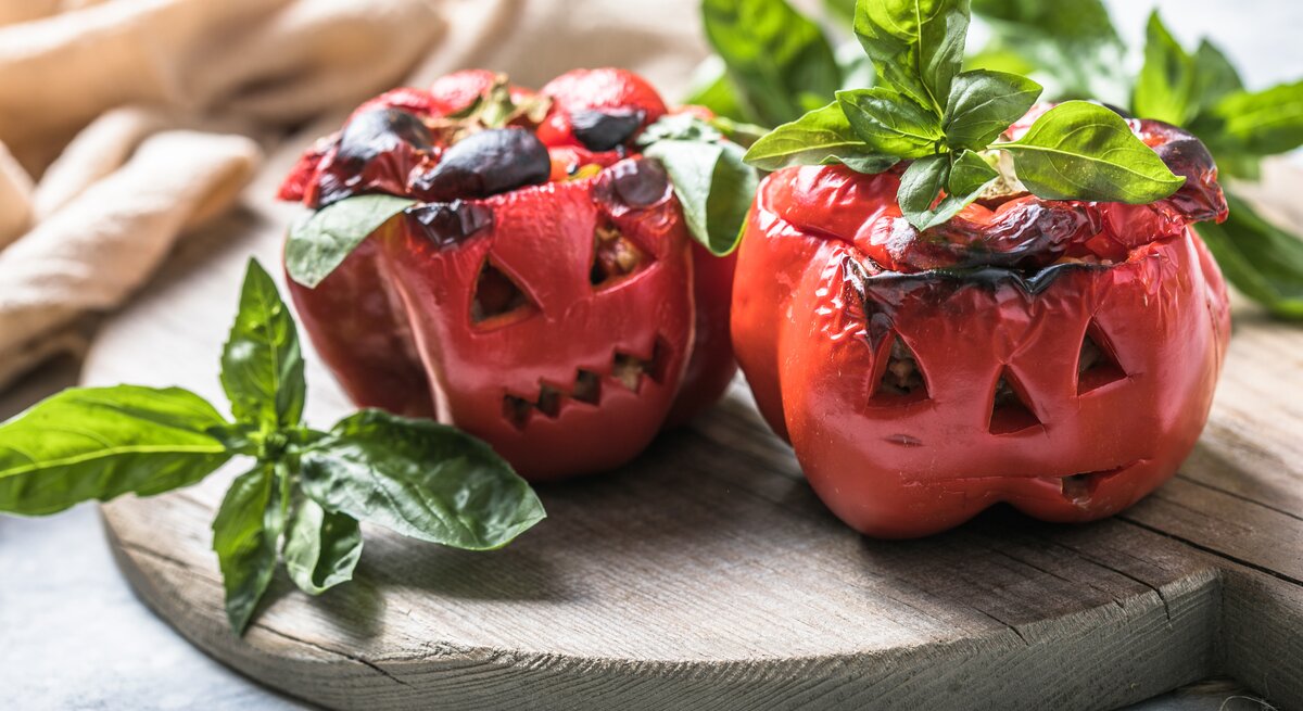 Zwei geschmorte Paprika im Halloween-Look auf einem Holzbrett mit Basilikum dekoriert. Alles von der Seite und recht nah fotografiert.