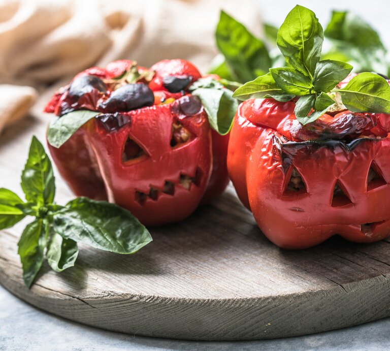 Zwei geschmorte Paprika im Halloween-Look auf einem Holzbrett mit Basilikum dekoriert. Alles von der Seite und recht nah fotografiert. | © AdobeStock/ Maria