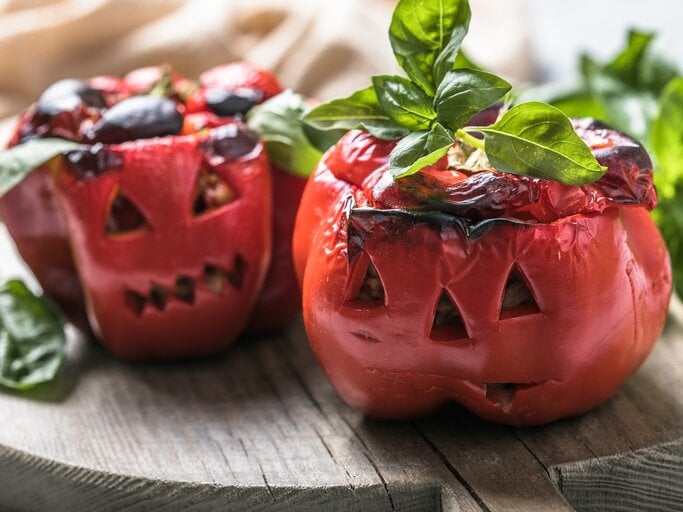 Zwei geschmorte Paprika im Halloween-Look auf einem Holzbrett mit Basilikum dekoriert. Alles von der Seite und recht nah fotografiert. | © AdobeStock/ Maria