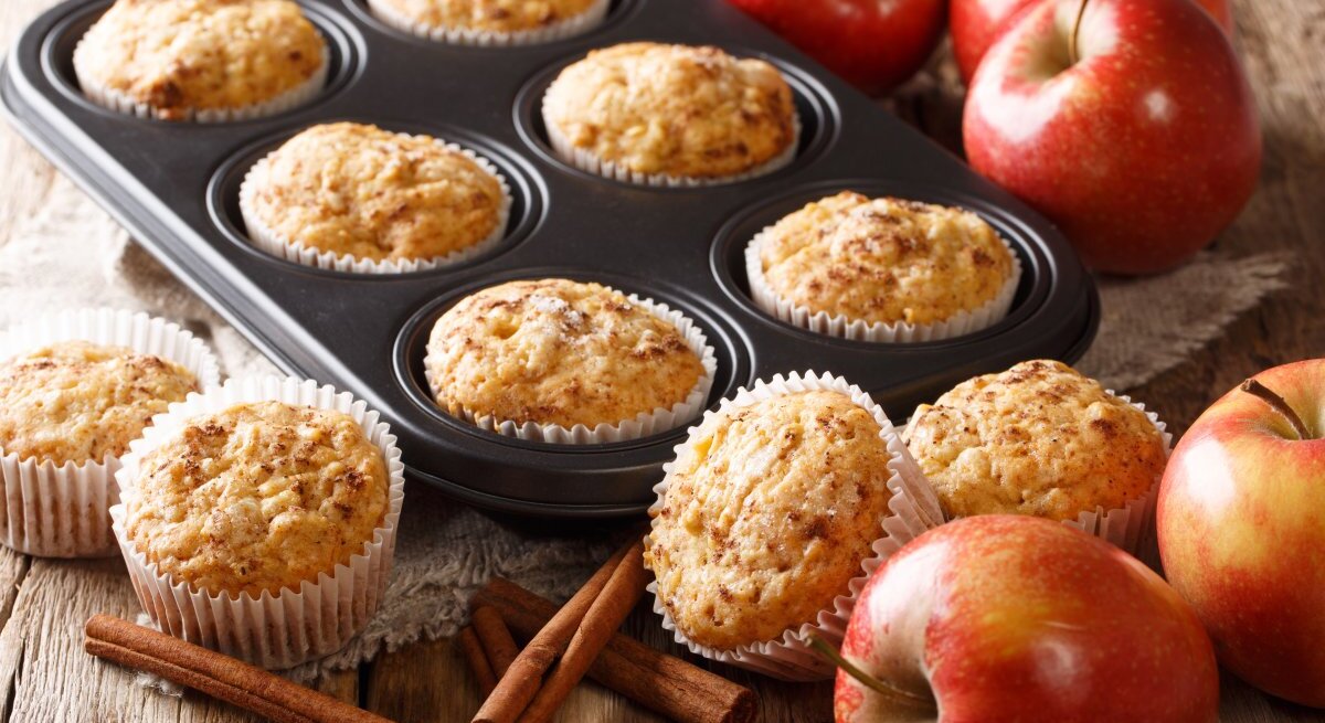 Frisch gebackene Apfelmuffins in einer Muffinform und auf einem Holztisch. Die Muffins sind goldbraun und sehen mit ihrer aufgegangenen, krümeligen Oberfläche appetitlich aus. Ein paar der Muffins sind noch in der schwarzen Muffinform, während andere auf dem Tisch neben der Form stehen, jeweils in weißen Papierförmchen. Rote Äpfel, Zimtstangen und ein Stück Sackleinen verleihen dem Bild eine rustikale und gemütliche Ausstrahlung. 