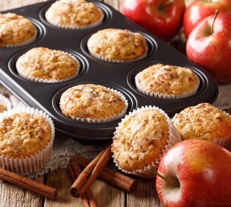 Frisch gebackene Apfelmuffins in einer Muffinform und auf einem Holztisch. Die Muffins sind goldbraun und sehen mit ihrer aufgegangenen, krümeligen Oberfläche appetitlich aus. Ein paar der Muffins sind noch in der schwarzen Muffinform, während andere auf dem Tisch neben der Form stehen, jeweils in weißen Papierförmchen. Rote Äpfel, Zimtstangen und ein Stück Sackleinen verleihen dem Bild eine rustikale und gemütliche Ausstrahlung.  | © Getty Images/ALLEKO