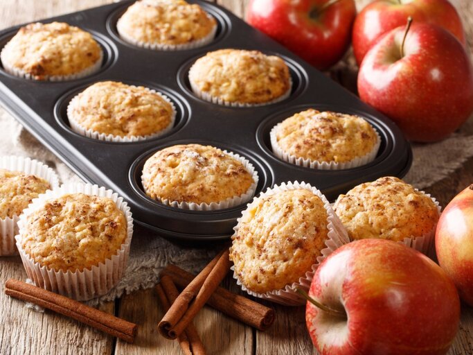 Frisch gebackene Apfelmuffins in einer Muffinform und auf einem Holztisch. Die Muffins sind goldbraun und sehen mit ihrer aufgegangenen, krümeligen Oberfläche appetitlich aus. Ein paar der Muffins sind noch in der schwarzen Muffinform, während andere auf dem Tisch neben der Form stehen, jeweils in weißen Papierförmchen. Rote Äpfel, Zimtstangen und ein Stück Sackleinen verleihen dem Bild eine rustikale und gemütliche Ausstrahlung.  | © Getty Images/ALLEKO