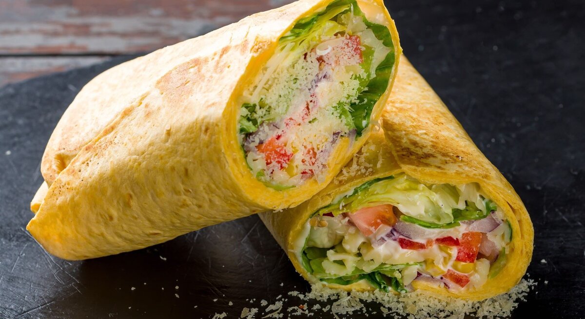 Zwei aufgeschnittene Tortillas gefüllt mit frischem Salat, Tomaten, Zwiebeln und Käse auf einer schwarzen Schieferplatte.