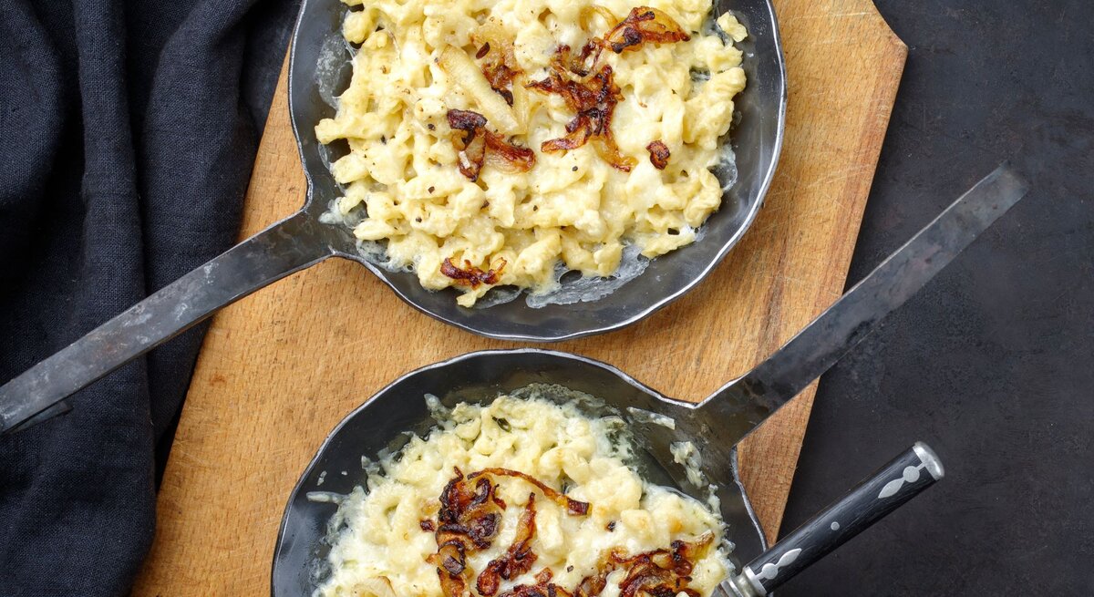 zwei gusseiserne Pfannen gefüllt mit Käsesätzle, garniert mit gebratenen Zwiebeln auf einer dunklen Tischdecke. 