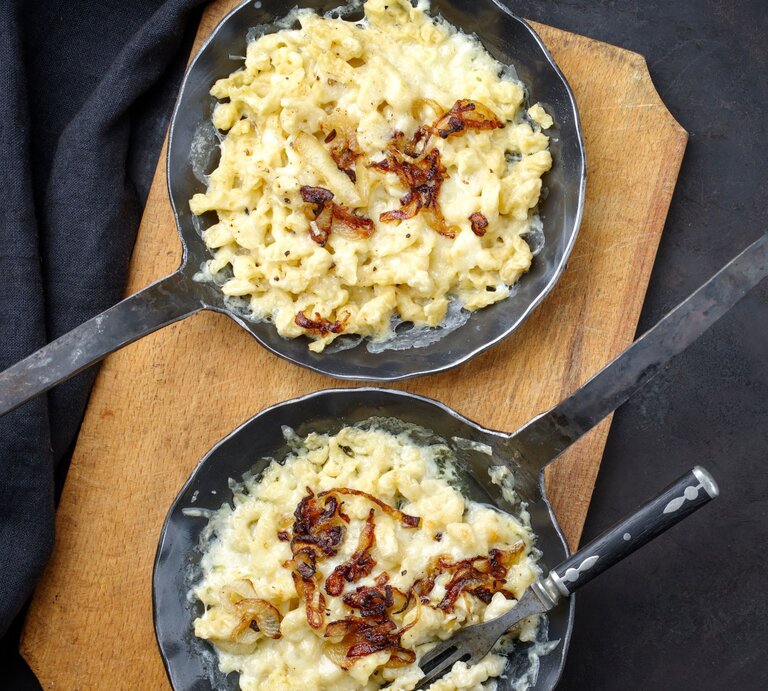 zwei gusseiserne Pfannen gefüllt mit Käsesätzle, garniert mit gebratenen Zwiebeln auf einer dunklen Tischdecke.  | © AdobeStock/ ABM