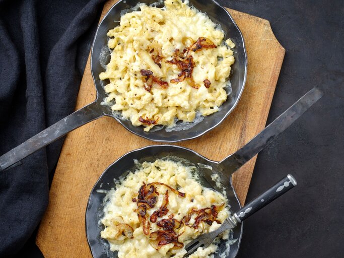 zwei gusseiserne Pfannen gefüllt mit Käsesätzle, garniert mit gebratenen Zwiebeln auf einer dunklen Tischdecke.  | © AdobeStock/ ABM