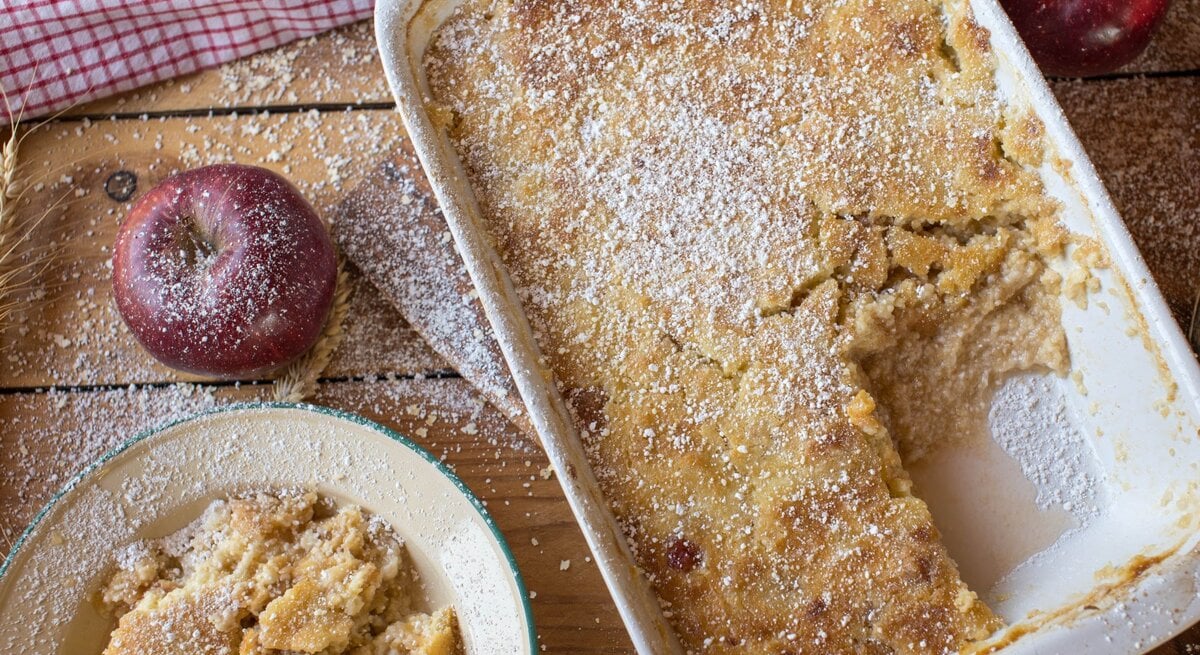 Frisch gebackener Apfelauflauf mit goldbrauner Kruste und Puderzucker in Auflaufform auf rustikalem Holztisch.