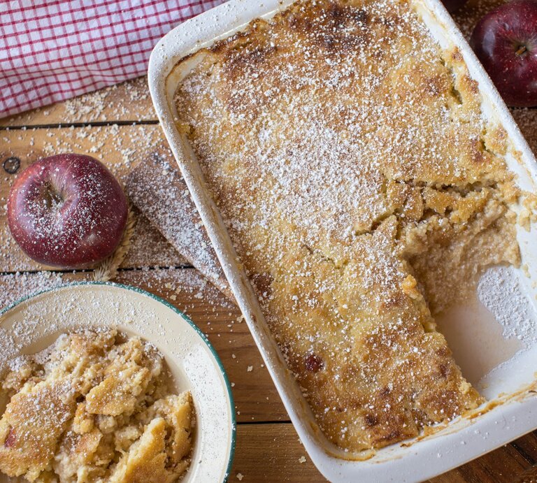 Frisch gebackener Apfelauflauf mit goldbrauner Kruste und Puderzucker in Auflaufform auf rustikalem Holztisch. | © Adobe Stock/ Angelika Heine
