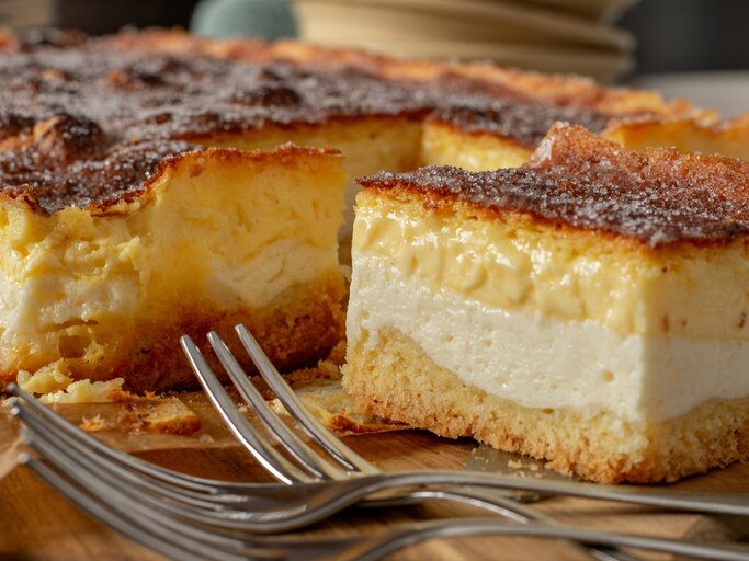 Der Blechkuchen mit einem Stück davon auf einem Teller und dunklen Schneidebrett nah fotografiert. | © Shutterstock/ Angelika Heine