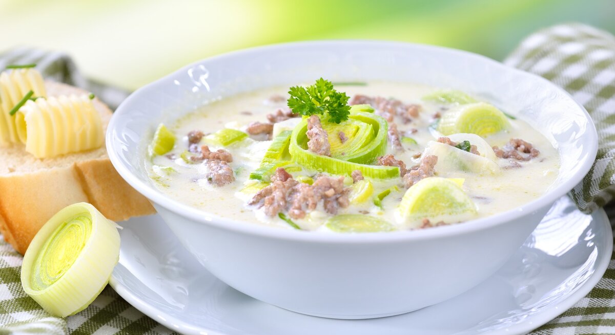 Das Bild zeigt eine Schüssel Suppe, die reichhaltig und cremig aussieht. Die Suppe ist mit Lauchringen garniert und es gibt sichtbare Stückchen von Hackfleisch. Neben der Suppenschüssel befindet sich eine Scheibe Brot mit einer Kugel Butter darauf. Das Brot und die Butter sehen frisch und einladend aus. Die Suppe scheint eine Käse-Lauch-Suppe zu sein, ein beliebtes Gericht in der deutschen Küche. 