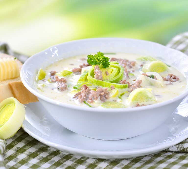 Das Bild zeigt eine Schüssel Suppe, die reichhaltig und cremig aussieht. Die Suppe ist mit Lauchringen garniert und es gibt sichtbare Stückchen von Hackfleisch. Neben der Suppenschüssel befindet sich eine Scheibe Brot mit einer Kugel Butter darauf. Das Brot und die Butter sehen frisch und einladend aus. Die Suppe scheint eine Käse-Lauch-Suppe zu sein, ein beliebtes Gericht in der deutschen Küche.  | © GettyImages/kabVisio