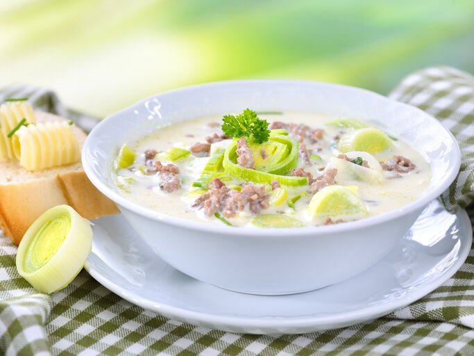 Das Bild zeigt eine Schüssel Suppe, die reichhaltig und cremig aussieht. Die Suppe ist mit Lauchringen garniert und es gibt sichtbare Stückchen von Hackfleisch. Neben der Suppenschüssel befindet sich eine Scheibe Brot mit einer Kugel Butter darauf. Das Brot und die Butter sehen frisch und einladend aus. Die Suppe scheint eine Käse-Lauch-Suppe zu sein, ein beliebtes Gericht in der deutschen Küche.  | © GettyImages/kabVisio