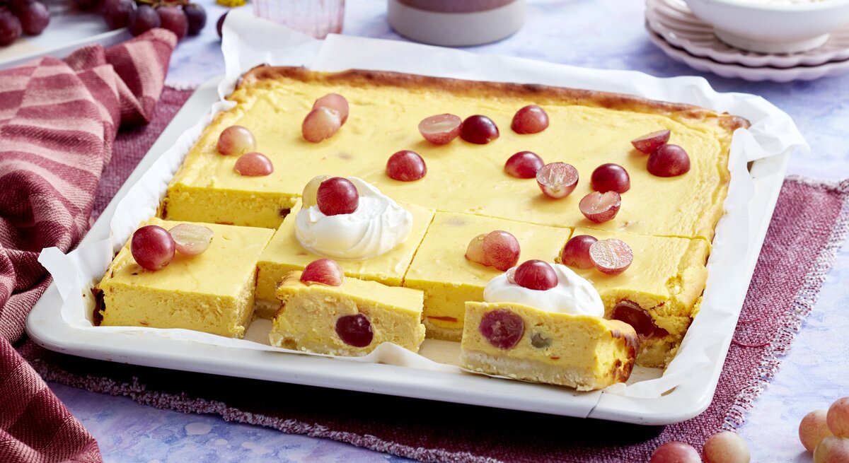 Ein weißes Blech mit dem Kürbis-Trauben-Cheesecake auf einem blau-weißem Marmortisch von schräg oben fotografiert