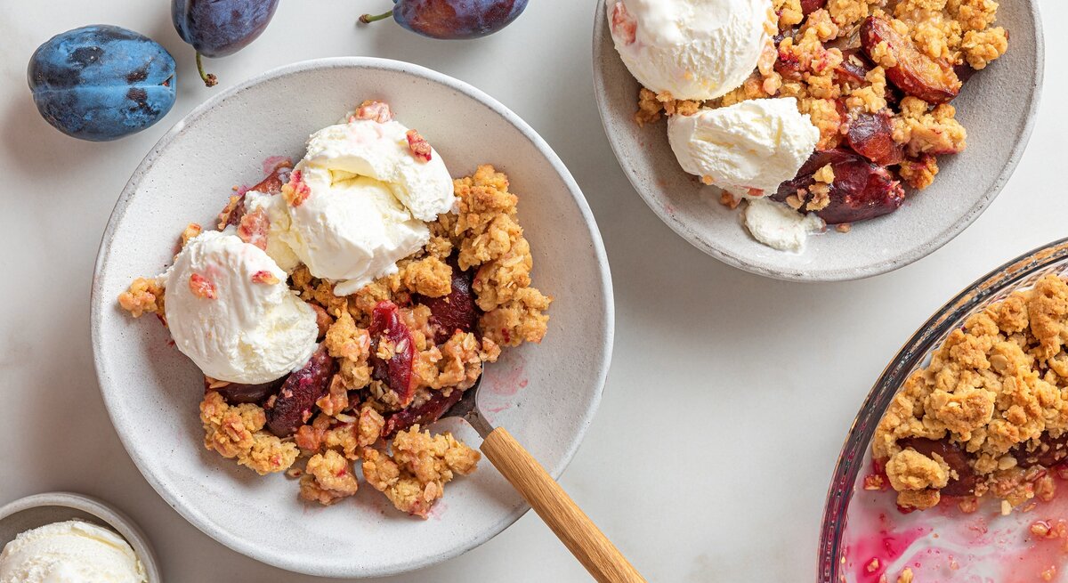 Zwei Teller mit dem Pflaumen-Crumble und Vanilleeis auf einem hellen Tisch mit weiteren frischen Pflaumen drum herum, direkt von oben fotografiert.
