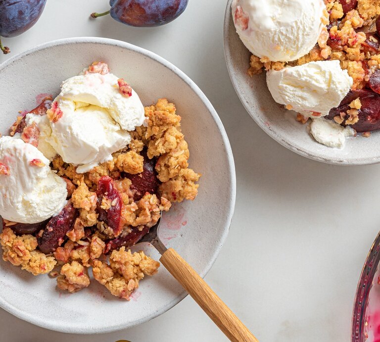Zwei Teller mit dem Pflaumen-Crumble und Vanilleeis auf einem hellen Tisch mit weiteren frischen Pflaumen drum herum, direkt von oben fotografiert. | © Shutterstock/ artem evdokimov