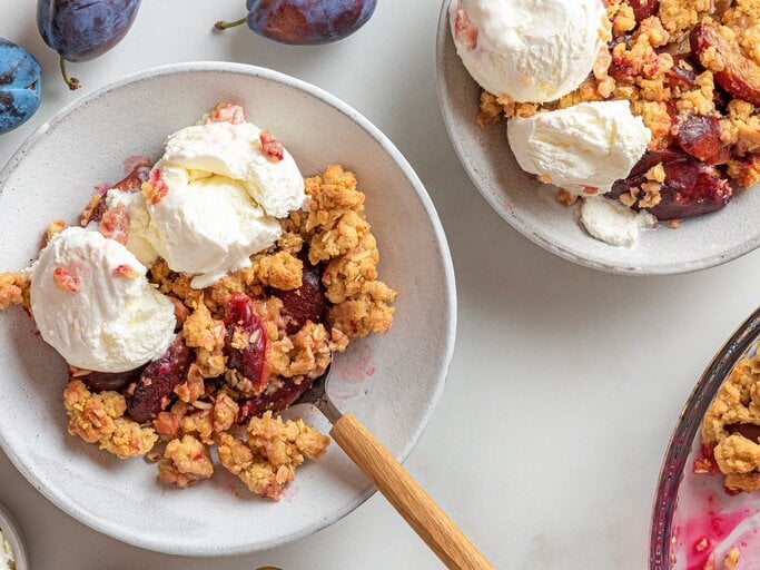 Zwei Teller mit dem Pflaumen-Crumble und Vanilleeis auf einem hellen Tisch mit weiteren frischen Pflaumen drum herum, direkt von oben fotografiert. | © Shutterstock/ artem evdokimov