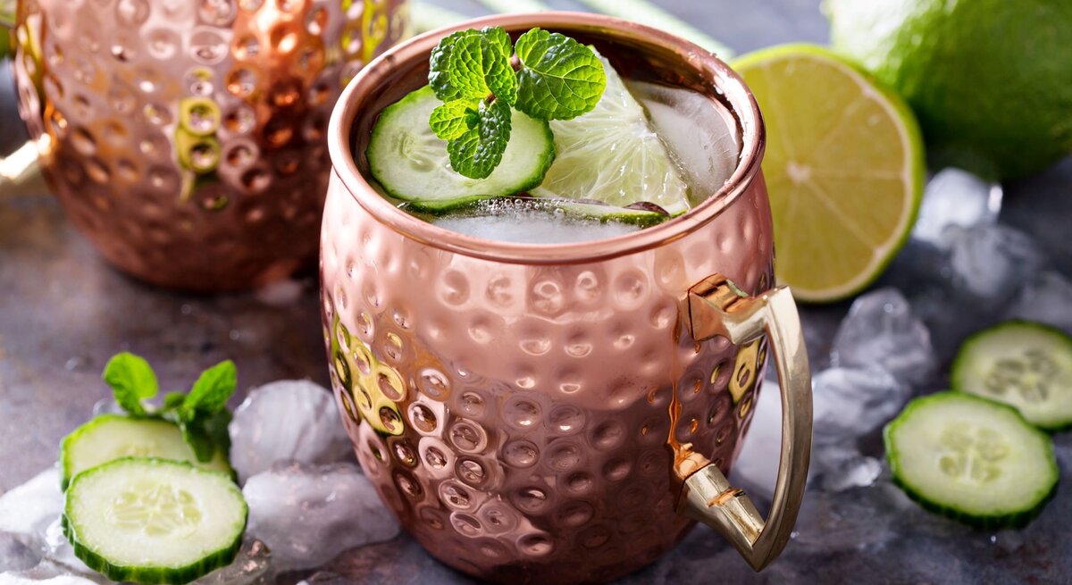 Moscow Mule in einem gehämmerten Kupferbecher mit Eis, Limettenscheibe, Gurke und Minze auf einem Tisch mit Eiswürfeln und Limetten