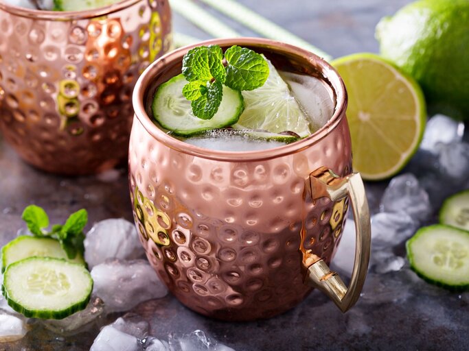 Moscow Mule in einem gehämmerten Kupferbecher mit Eis, Limettenscheibe, Gurke und Minze auf einem Tisch mit Eiswürfeln und Limetten | © AdobeStock/ fahrwasser