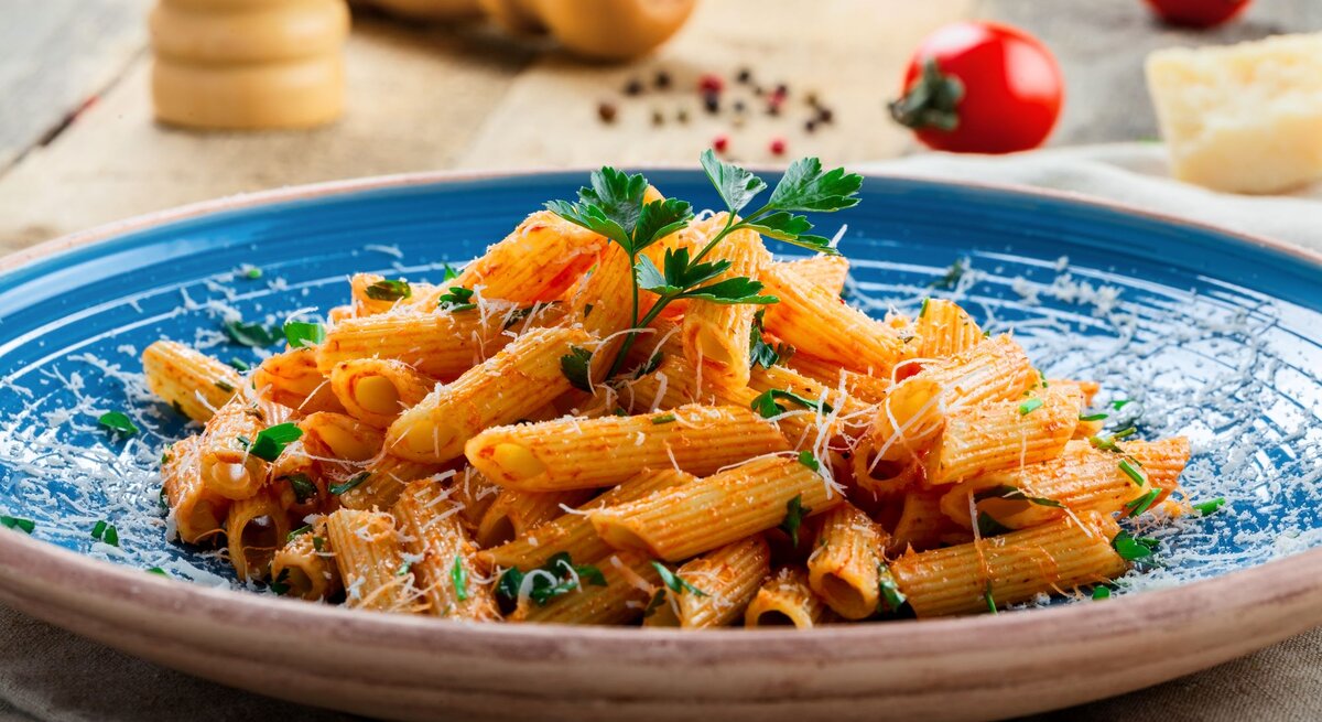 Penne in leuchtender Tomatensauce mit Parmesan und Kräutern serviert auf einem blauen Teller vor rustikalem Hintergrund.