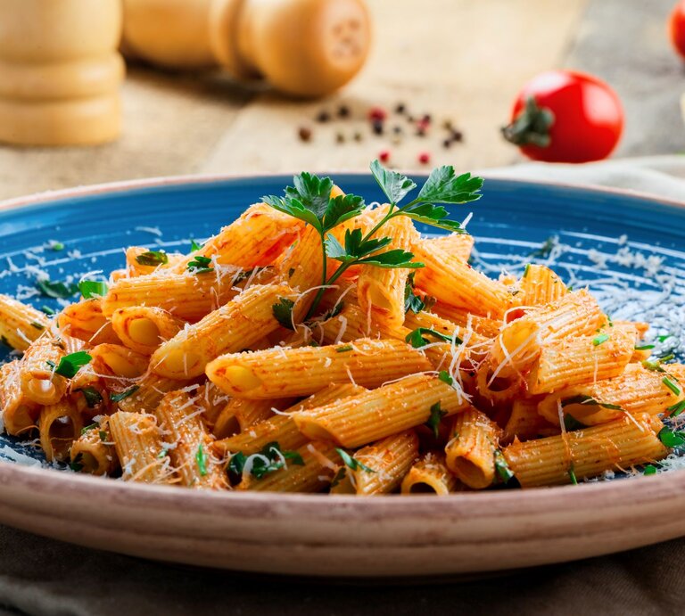 Penne in leuchtender Tomatensauce mit Parmesan und Kräutern serviert auf einem blauen Teller vor rustikalem Hintergrund. | © AdobeStock/Maxim Khytra