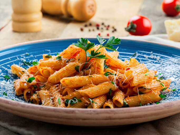 Penne in leuchtender Tomatensauce mit Parmesan und Kräutern serviert auf einem blauen Teller vor rustikalem Hintergrund. | © AdobeStock/Maxim Khytra