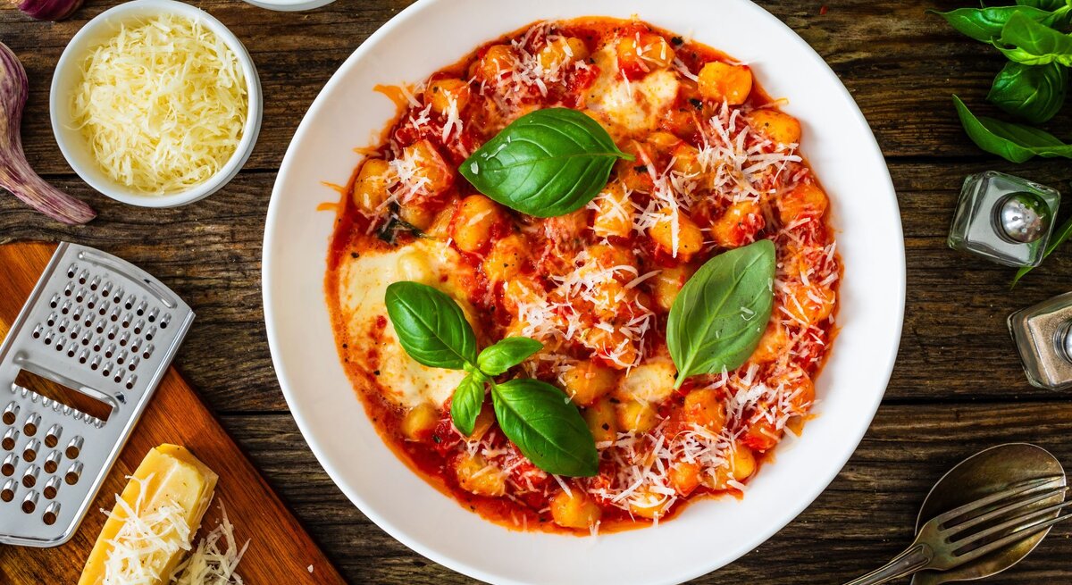 Frisch zubereitete Gnocchi in Tomatensauce mit Basilikumblättern und geriebenem Parmesan auf rustikalem Holztisch.