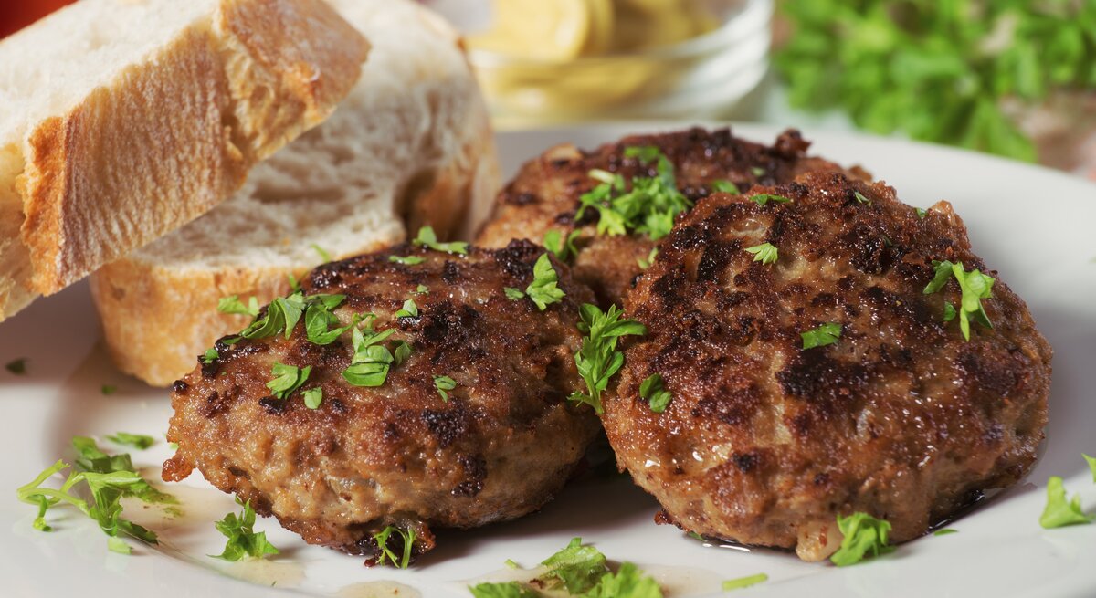 Mehrere Frikadellen bestreut mit Kräutern auf weißem Teller neben Brotscheiben. 