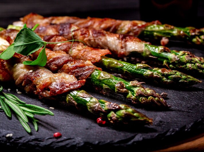 Appetitlicher Blick auf gebratenen Spargel in Speckmantel, fein gewürzt mit Kräutern auf Steinplatte. | © Shutterstock/sweet marshmallow