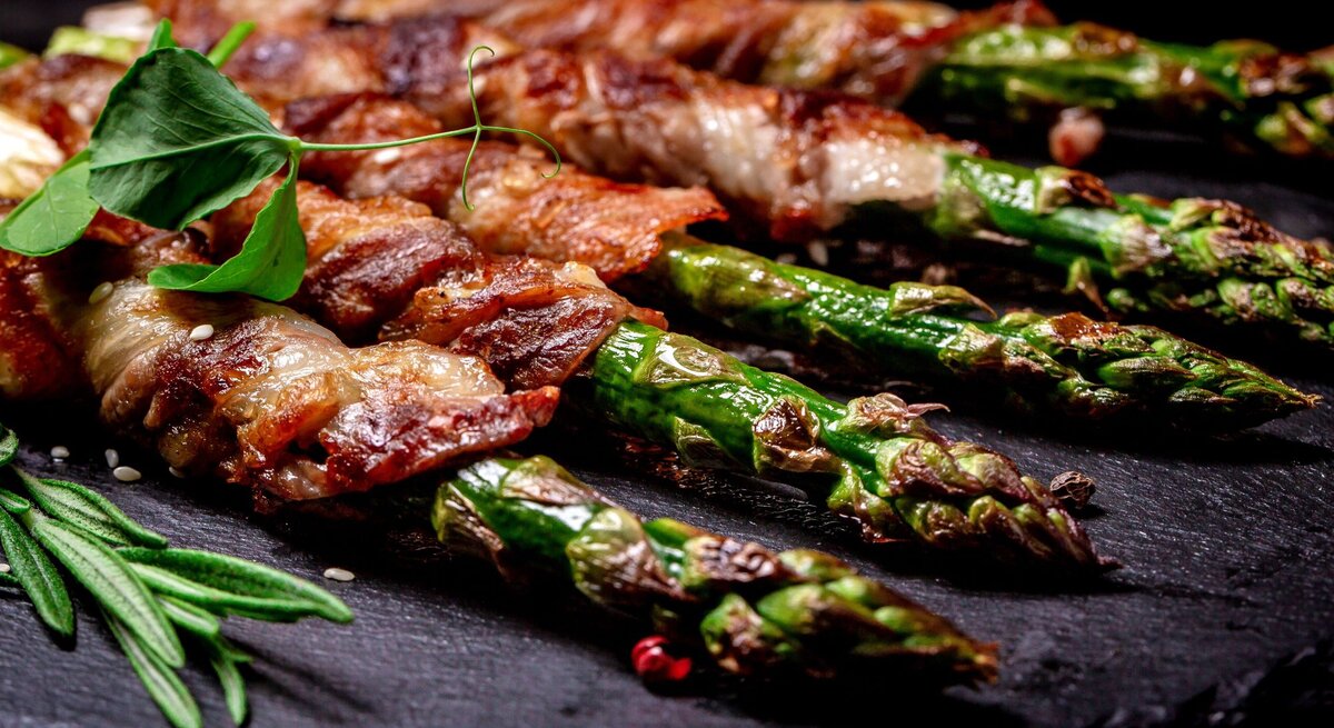 Appetitlicher Blick auf gebratenen Spargel in Speckmantel, fein gewürzt mit Kräutern auf Steinplatte.