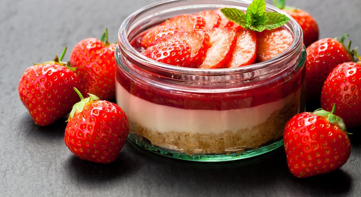 Frisch zubereitetes Erdbeerdessert im Glas mit Keksboden, Sahneschicht und Erdbeerscheiben, umgeben von frischen Erdbeeren auf dunklem Hintergrund.