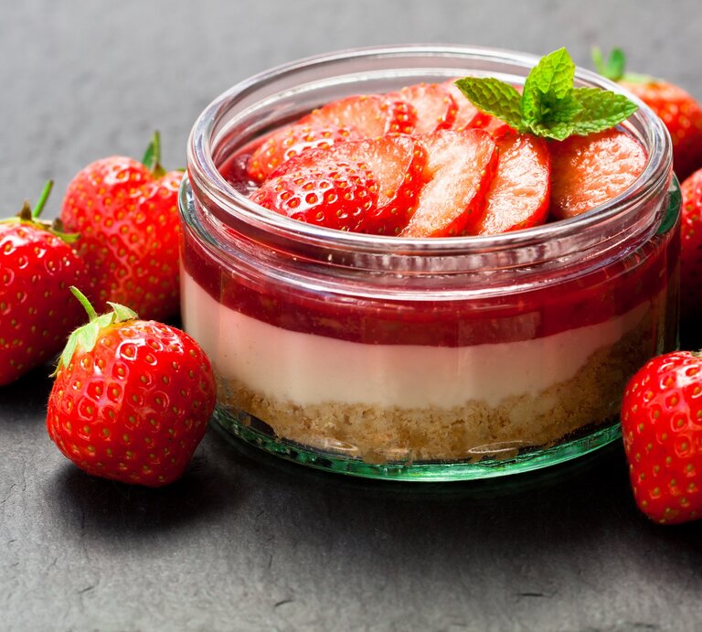 Frisch zubereitetes Erdbeerdessert im Glas mit Keksboden, Sahneschicht und Erdbeerscheiben, umgeben von frischen Erdbeeren auf dunklem Hintergrund. | © iStock/Lena_Zajchikova
