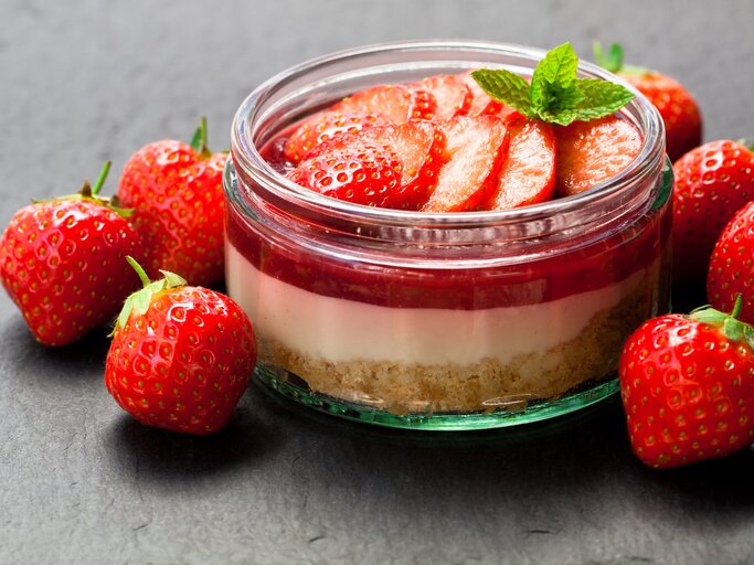 Frisch zubereitetes Erdbeerdessert im Glas mit Keksboden, Sahneschicht und Erdbeerscheiben, umgeben von frischen Erdbeeren auf dunklem Hintergrund. | © iStock/Lena_Zajchikova