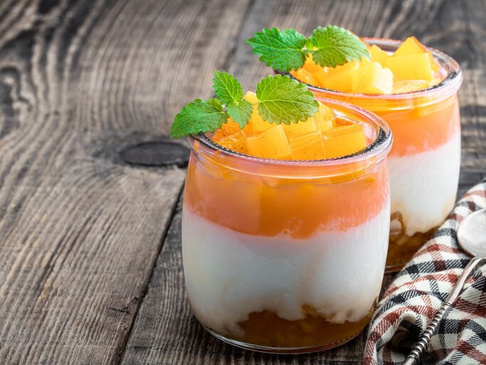 Solero-Dessert im Glas, mit frischen Pfirsichstücken und Minze dekoriert, auf einem rustikalen Holztisch angerichtet. | © Getty Images/Lena_Zajchikova
